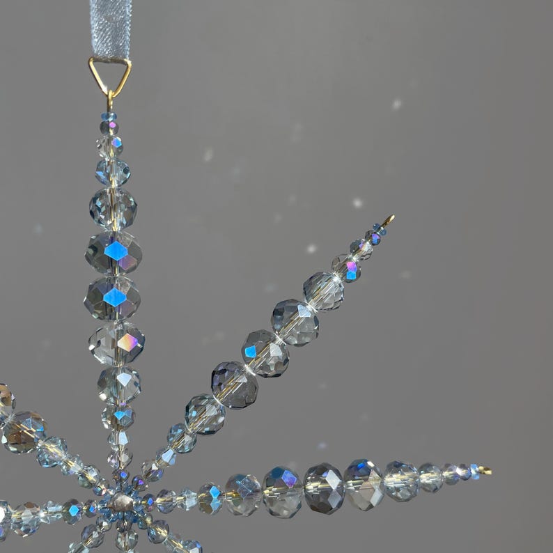 Pode incluir: Um ornamento em forma de floco de neve constru&iacute;do com contas de cristal facetadas, transl&uacute;cidas, cinzentas e iridescentes. A estrutura apresenta v&aacute;rios bra&ccedil;os radiantes de tamanhos variados, ligados por fio dourado e suspensos por uma fita azul clara.