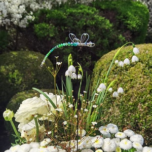 Deko-Libelle auf Steckstab steckt zur Zierde in einem Blumen bouquet.
Im Hintergrund sieht man den Garten.