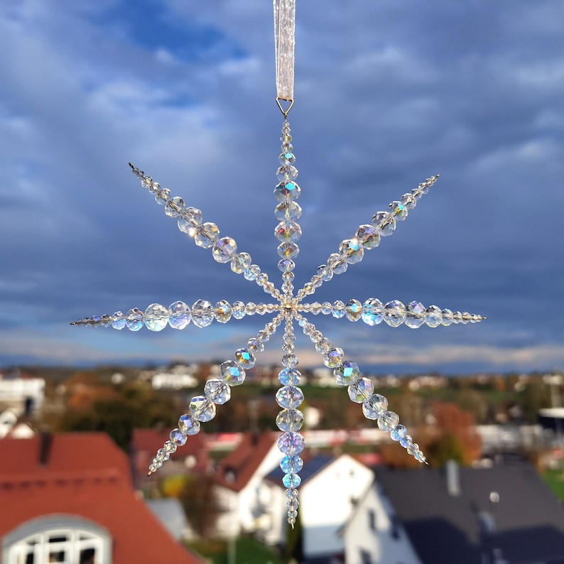 Ein Stern aus glitzernden, transparenten Kristallperlen hängt an einem Organzaband.
Im Hintergrund sieht man eine Landschaft mit Häusern und blauem Himmel.