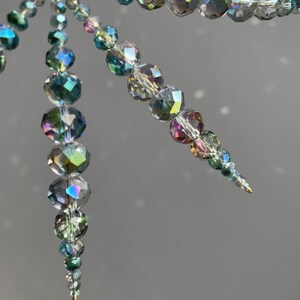 Pode incluir: Um ornamento decorativo suspenso composto por m&uacute;ltiplos fios de contas de vidro facetadas e iridescentes. As contas apresentam um espectro de cores incluindo azul, verde e prata, dispostas num design c&oacute;nico que estreita em dire&ccedil;&atilde;o &agrave; base.