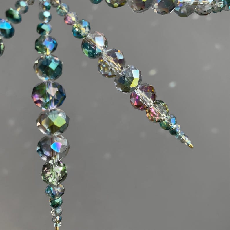 Pode incluir: Um ornamento decorativo suspenso composto por m&uacute;ltiplos fios de contas de vidro facetadas e iridescentes. As contas apresentam um espectro de cores incluindo azul, verde e prata, dispostas num design c&oacute;nico que estreita em dire&ccedil;&atilde;o &agrave; base.