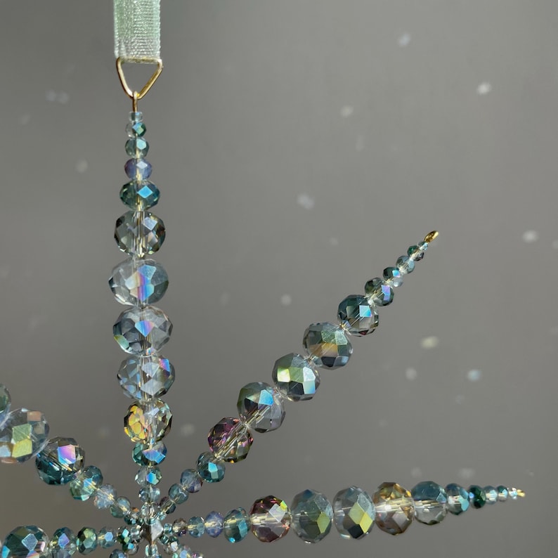 Pode incluir: Um ornamento em forma de estrela constru&iacute;do com contas de vidro facetadas e iridescentes em tons de azul, cinza e prata. As contas est&atilde;o dispostas em pontas radiantes que diminuem de tamanho em dire&ccedil;&atilde;o &agrave;s pontas, suspenso por uma fita verde clara.