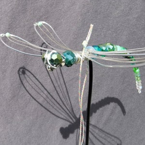 Libellen Dekostecker aus grünen, facettierten Rondell-Perlen aus Glas vor grauem Hintergrund. Die Flügel sind aus Draht geformt.
Die Perlenfunkeln in der Sonne.