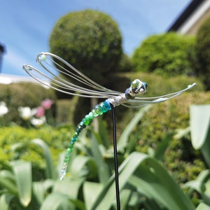 Grüne Deko-Libelle auf Steckstab steckt im Blumenbeet eines Gartens.