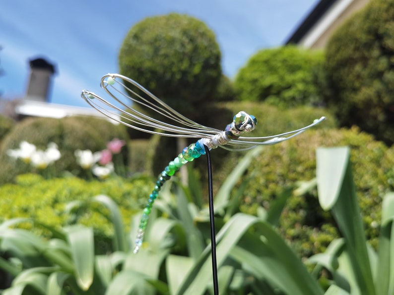 Grüne Deko-Libelle auf Steckstab steckt im Blumenbeet eines Gartens.