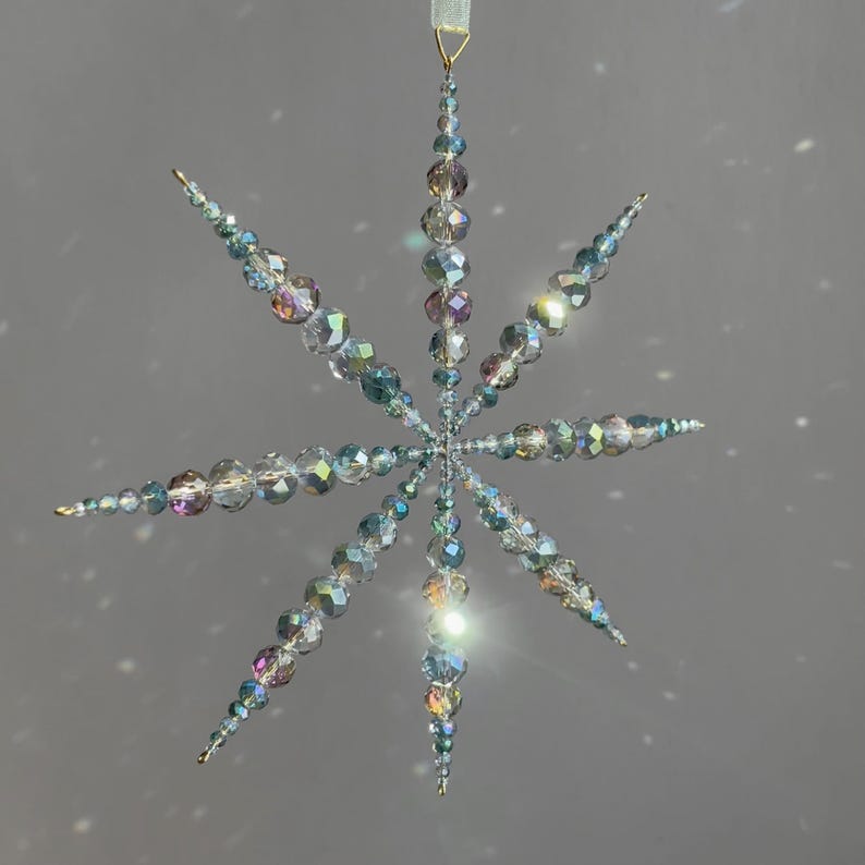 Pode incluir: Um ornamento suspenso em forma de estrela feito de contas de vidro facetadas e iridescentes. O design apresenta oito pontas radiantes com contas de tamanhos variados, criando uma apar&ecirc;ncia cristalina e reflexiva com tons subtis de arco-&iacute;ris.