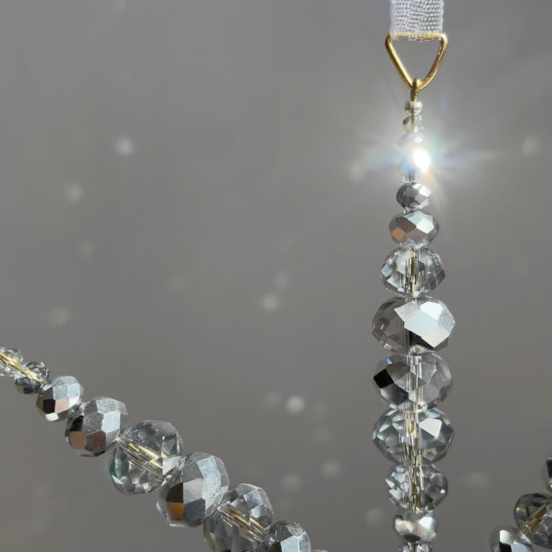 Pode incluir: Um ornamento decorativo suspenso com m&uacute;ltiplos fios de contas facetadas na cor prata met&aacute;lica. As contas est&atilde;o dispostas em v&aacute;rios tamanhos e formas, suspensas por um aro met&aacute;lico triangular de tom dourado com uma fita branca.