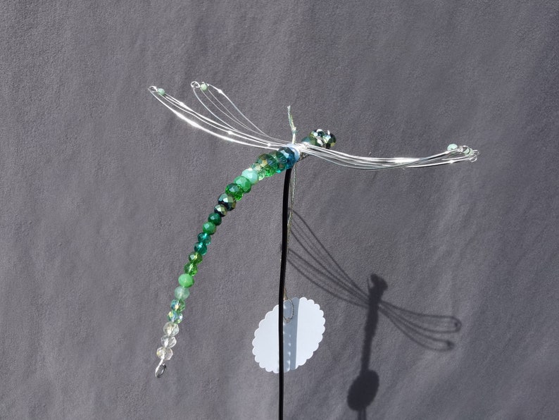 Libellen Pflanzenstecker aus grünen, facettierten Rondell-Perlen aus Glas vor grauem Hintergrund. Die Flügel sind aus Draht geformt.
Die Perlenfunkeln in der Sonne.