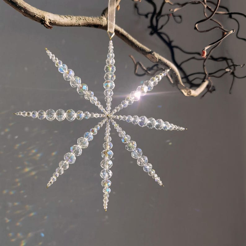 Pode incluir: Um ornamento em forma de estrela constru&iacute;do com contas de cristal transparente facetado dispostas em oito pontas radiantes. As contas variam em tamanho, estreitando-se em dire&ccedil;&atilde;o &agrave;s extremidades de cada bra&ccedil;o. Um la&ccedil;o de fita clara est&aacute; preso.