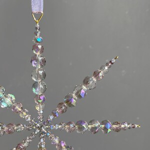 Pode incluir: Um ornamento em forma de estrela feito de contas de vidro facetadas e iridescentes em tons transparentes e roxo claro. As contas est&atilde;o dispostas em pontas radiantes, ligadas por fio dourado e suspensas por uma fita roxa clara no topo.