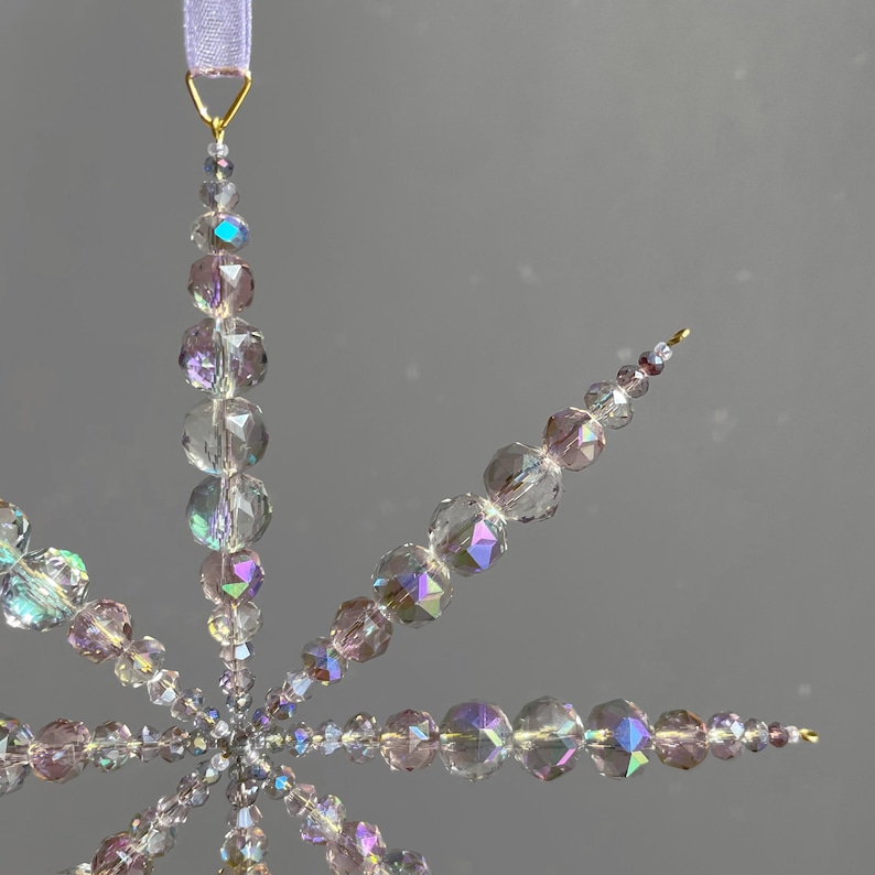 Pode incluir: Um ornamento em forma de estrela feito de contas de vidro facetadas e iridescentes em tons transparentes e roxo claro. As contas est&atilde;o dispostas em pontas radiantes, ligadas por fio dourado e suspensas por uma fita roxa clara no topo.