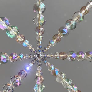 Pode incluir: Um ornamento em forma de estrela constru&iacute;do com contas de vidro facetadas e iridescentes. As contas est&atilde;o dispostas em linhas radiantes que se estendem a partir de um ponto central, apresentando um espectro de cores que refletem a luz, incluindo tons claros, rosa e azul.