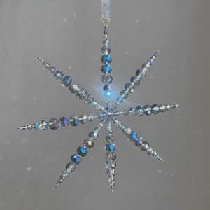 Pode incluir: Um ornamento suspenso em forma de estrela feito de contas de vidro facetadas e iridescentes, transparentes e azul-claras. O ornamento apresenta oito pontas radiantes com contas de v&aacute;rios tamanhos, ligadas por um centro e suspensas por uma fita azul-clara.