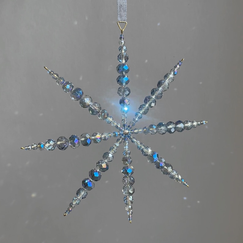 Pode incluir: Um ornamento suspenso em forma de estrela feito de contas de vidro facetadas e iridescentes, transparentes e azul-claras. O ornamento apresenta oito pontas radiantes com contas de v&aacute;rios tamanhos, ligadas por um centro e suspensas por uma fita azul-clara.
