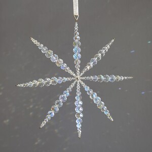 Pode incluir: Um ornamento suspenso em forma de estrela constru&iacute;do com contas de cristal transparente facetadas. O design apresenta oito pontas radiantes com contas de tamanhos variados, criando um padr&atilde;o geom&eacute;trico. Uma fita branca est&aacute; presa na ponta superior.
