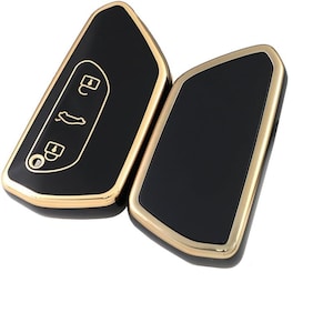 May include: Two black car key fobs with gold-colored trim. One fob has white icons for lock, unlock, and trunk release. The other fob is solid black with a gold-colored trim. The fobs are on a white background.