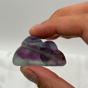 May include: A carved fluorite cloud sculpture. The stone features a gradient of colors, including purple, lavender, and clear. The cloud shape is smooth and polished, with rounded edges. The sculpture is held in a person's hand.