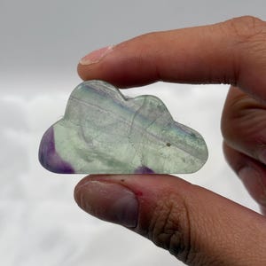 May include: A cloud-shaped crystal carving. The translucent stone features swirls of purple, green, and clear hues. The polished surface reflects light, showcasing the natural patterns within the crystal. A unique decorative item.