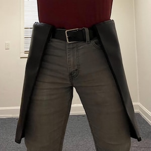 May include: Black leather chaps worn over gray jeans, secured with a black belt and silver buckle. The chaps extend down the legs, creating a distinct silhouette. The person is wearing a maroon shirt.