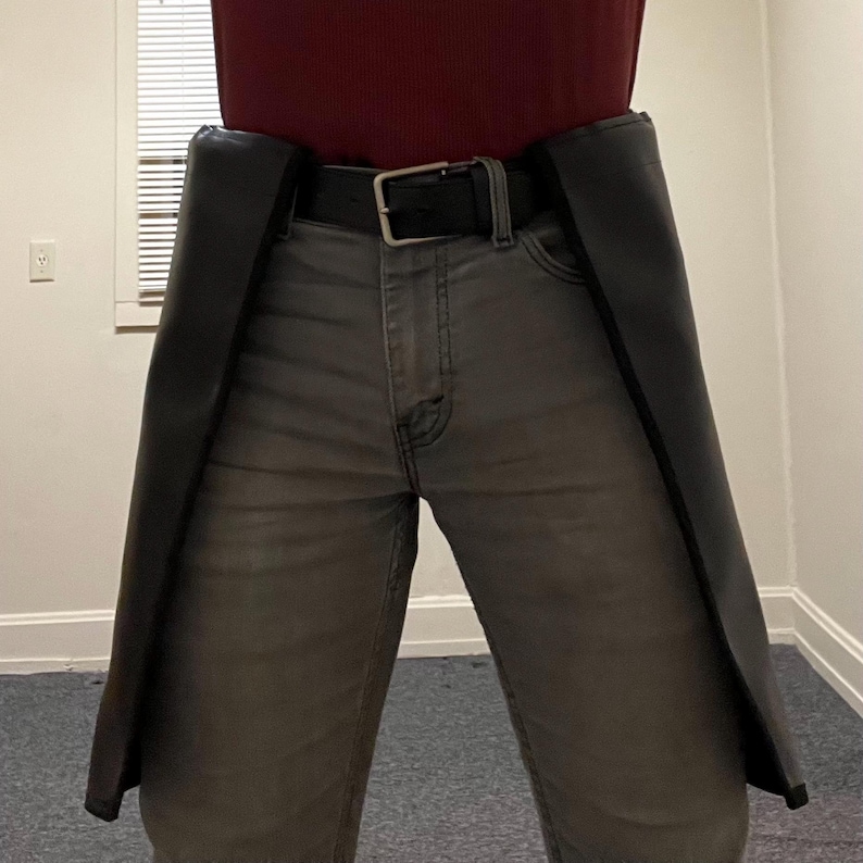 May include: Black leather chaps worn over gray jeans, secured with a black belt and silver buckle. The chaps extend down the legs, creating a distinct silhouette. The person is wearing a maroon shirt.