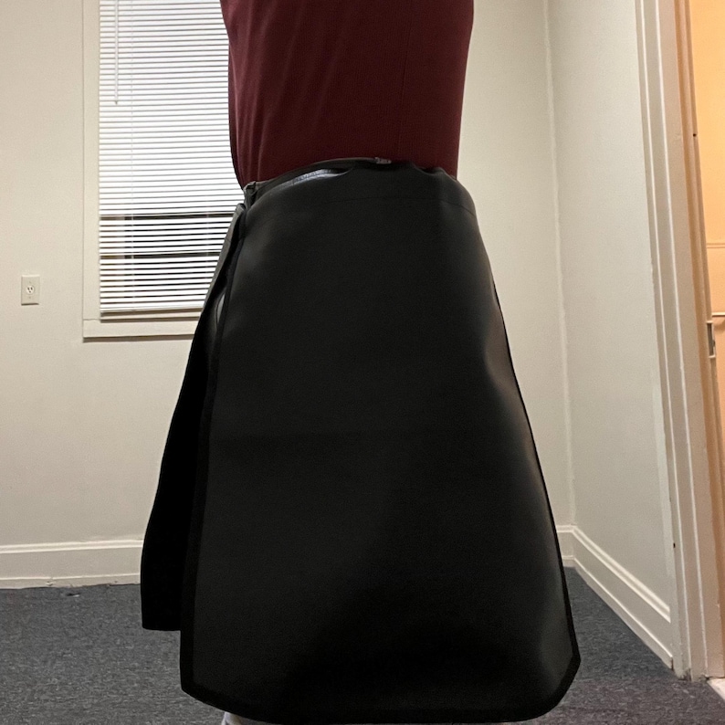 May include: Black wrap skirt made of a smooth material. The skirt has a simple design with a wrap-around style and a slight A-line shape. The skirt is shown from the waist down.