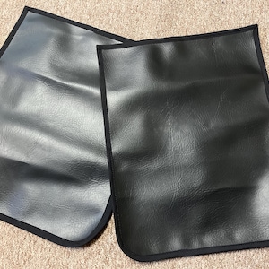 May include: Two black rectangular leather-like mats with rounded corners and a black trim. The mats are placed side by side, with a textured surface. The background is a light brown, possibly carpeted surface.