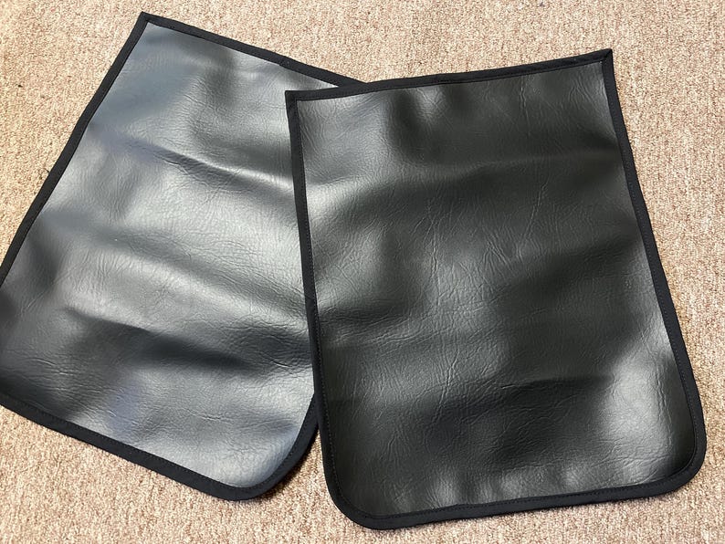 May include: Two black rectangular leather-like mats with rounded corners and a black trim. The mats are placed side by side, with a textured surface. The background is a light brown, possibly carpeted surface.