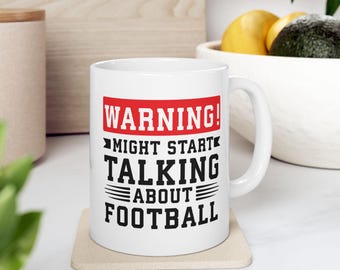 May include: White ceramic mug with the text "WARNING! MIGHT START TALKING ABOUT FOOTBALL" in black and red. The mug is on a beige coaster. The handle is on the right side of the mug.