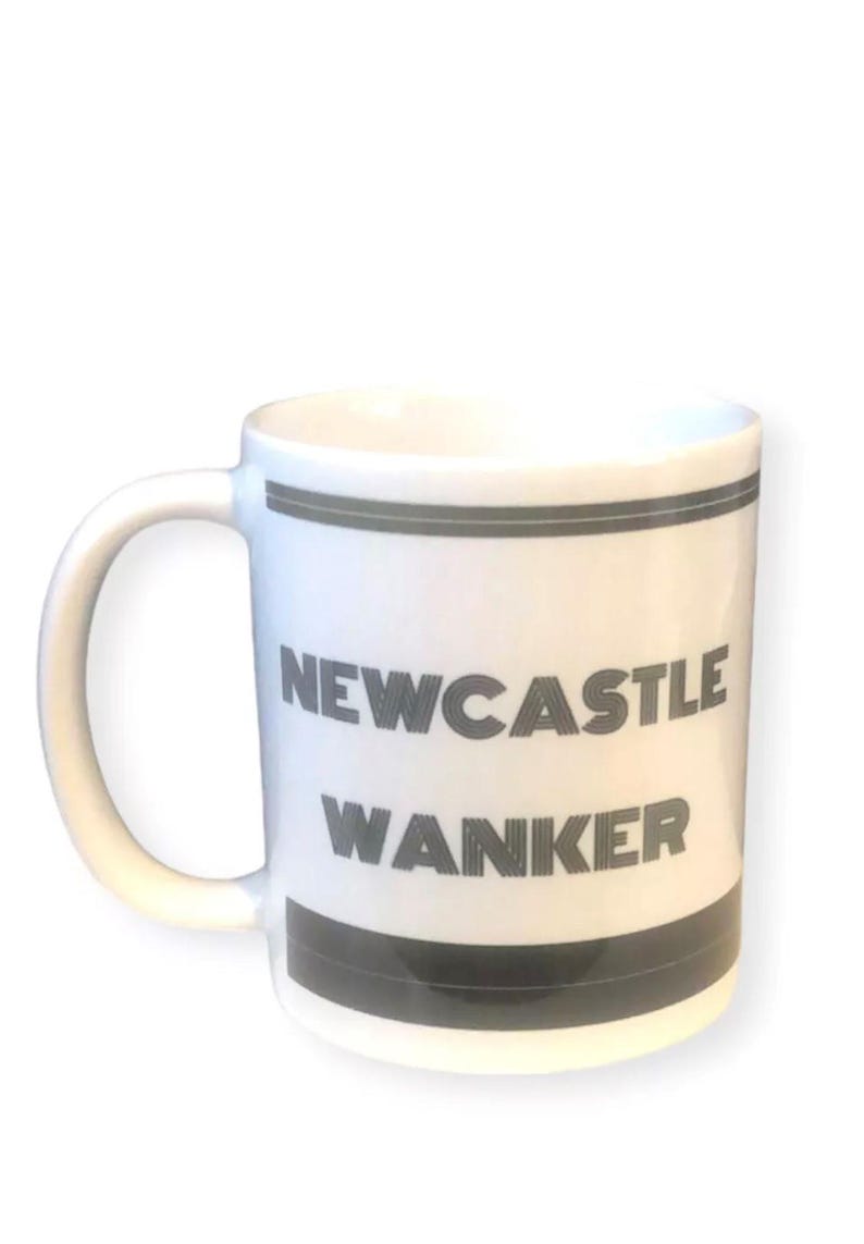 May include: White ceramic mug with a curved handle. The mug features the text "NEWCASTLE WANKER" in gray block letters, with gray bands at the top and bottom. The mug is set against a white background.