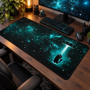 May include: A rectangular desk mat featuring a deep space galaxy print in shades of black and teal. The design includes a graphic of a black cat silhouette being lifted by a bright white beam of light from a circular flying saucer above.
