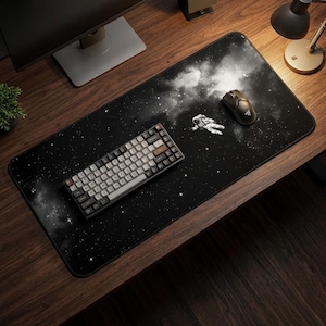 May include: A black desk mat featuring a starry space design with an astronaut illustration. A keyboard and a computer mouse are on the mat. The keyboard has white and gray keys. The mouse is black.
