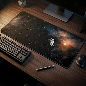 May include: A large rectangular mousepad featuring a cosmic design with a starry night sky and an astronaut. The pad has a black border and is placed on a wooden desk next to a keyboard, mouse, and pen.