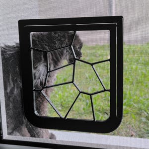 May include: A black pet door installed in a screen window. The door has a decorative geometric design and allows a view of the green grass outside. A fluffy, dark-colored cat is visible through the screen.