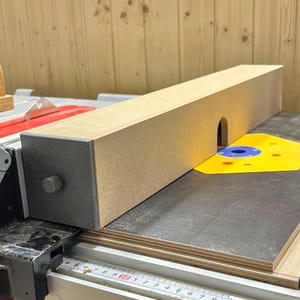 Puede incluir: Un accesorio de sierra de mesa de madera con forma rectangular y una pieza final de color gris oscuro. El accesorio tiene una pequeña abertura y está colocado sobre una hoja de sierra. La hoja de sierra es plateada.