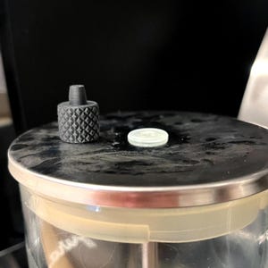 Puede incluir: Primer plano de un botón gris oscuro texturizado y un pequeño disco blanco sobre un recipiente de acero inoxidable. El botón tiene un patrón de diamantes. El recipiente tiene un borde de plástico transparente.