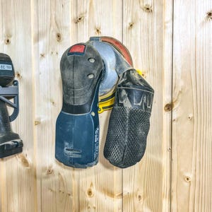 May include: A cordless random orbital sander with a black and blue body, red power button, and a silver sanding disc. A black mesh dust bag is attached. The sander is mounted on a light wood panel wall.