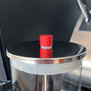 May include: A red bottle stopper with a diamond pattern sits atop a glass container. The stopper is cylindrical with a white band separating the top and bottom sections. The container has a silver rim.