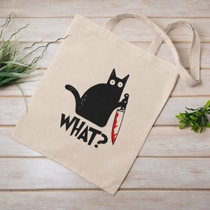 Op de afbeelding: Beige canvas draagtas met een zwarte kattenafbeelding die een bloedig mes vasthoudt. Het woord "WHAT?" staat eronder gedrukt. De tas heeft twee beige handvatten en ligt op een licht houten oppervlak.