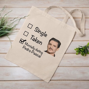Puede incluir: Bolso tote de lona beige con el texto "Single", "Taken" y "Mentally dating Pedro Pascal" con una marca de verificación junto a "Taken". La bolsa presenta una imagen impresa de Pedro Pascal.
