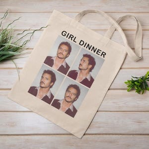 Peut inclure: Un sac fourre-tout beige avec le texte "GIRL DINNER" et quatre images d'un homme. Le sac a deux poignées et est posé sur une surface en bois clair. Des plantes vertes sont à gauche et à droite.