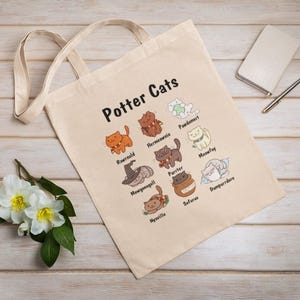 Könnte beinhalten: Beige Tragetasche mit dem Text "Potter Cats" und Illustrationen von Katzen in verschiedenen Posen. Die Tasche hat zwei Griffe und besteht aus einem leinwandartigen Material. Ein Notizbuch, ein Stift und Blumen sind ebenfalls im Bild.