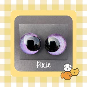 May include: Two round, iridescent purple and black earrings. The earrings have a glittery, speckled appearance with a black center. The word "Pixie" is written in white script below the earrings.