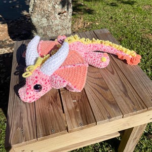 May include: A handmade crochet dragon in shades of pink, peach, and yellow, with white horns and large, glittery eyes. The dragon is lying on a wooden surface, showcasing its detailed craftsmanship and soft texture.