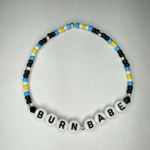 May include: A white beaded necklace with black and blue beads. The necklace spells out "BURN BABE" in black letters on white beads.