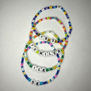 Könnte beinhalten: Eine Sammlung von Armbändern mit bunten Perlen. Einige Armbänder haben weiße Buchstabenperlen mit der Aufschrift "1993", "10 MILER", "10K" und "5K". Die Armbänder bestehen aus kleinen, farbigen Perlen.