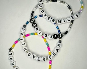 May include: Four beaded bracelets with positive messages: "You Got This", "Faith Over Fear", "Don't Quit", and "Wind Over Miles". The bracelets are made with white beads and black lettering. The bracelets are on a white background.