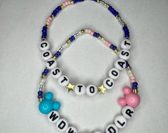 Puede incluir: Dos pulseras de cuentas. La pulsera superior tiene el texto "COAST TO COAST" con cuentas de estrella doradas. La pulsera inferior tiene el texto "WDW TODLR" con una cabeza de Mickey Mouse azul. Las pulseras tienen cuentas azules, rosas y blancas.