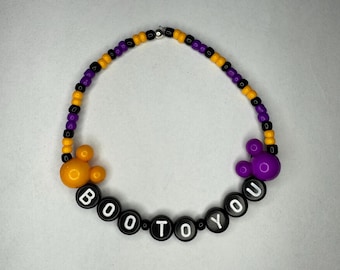 May include: A black beaded necklace with purple and yellow beads. The necklace has the words "BOO TO YOU" spelled out in black beads and features purple and yellow Mickey Mouse ears.