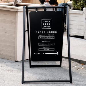 May include: A black A-frame sidewalk sign with white text reading Daily Dose Coffee, Store Hours, Monday-Friday 8:00am-6:00pm, Saturday 10:00am-11:00pm, Sunday Closed, and Come In with an arrow pointing to the right, mounted on a black metal frame.