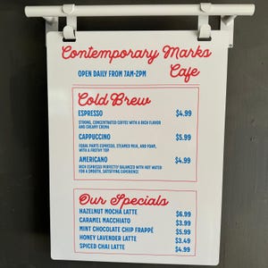 May include: White sign with red and blue text listing the menu for Contemporary Marks Cafe. The menu includes cold brew coffee options like espresso, cappuccino, and Americano, as well as specialty drinks like hazelnut mocha latte, caramel macchiato, and mint chocolate chip frappe.